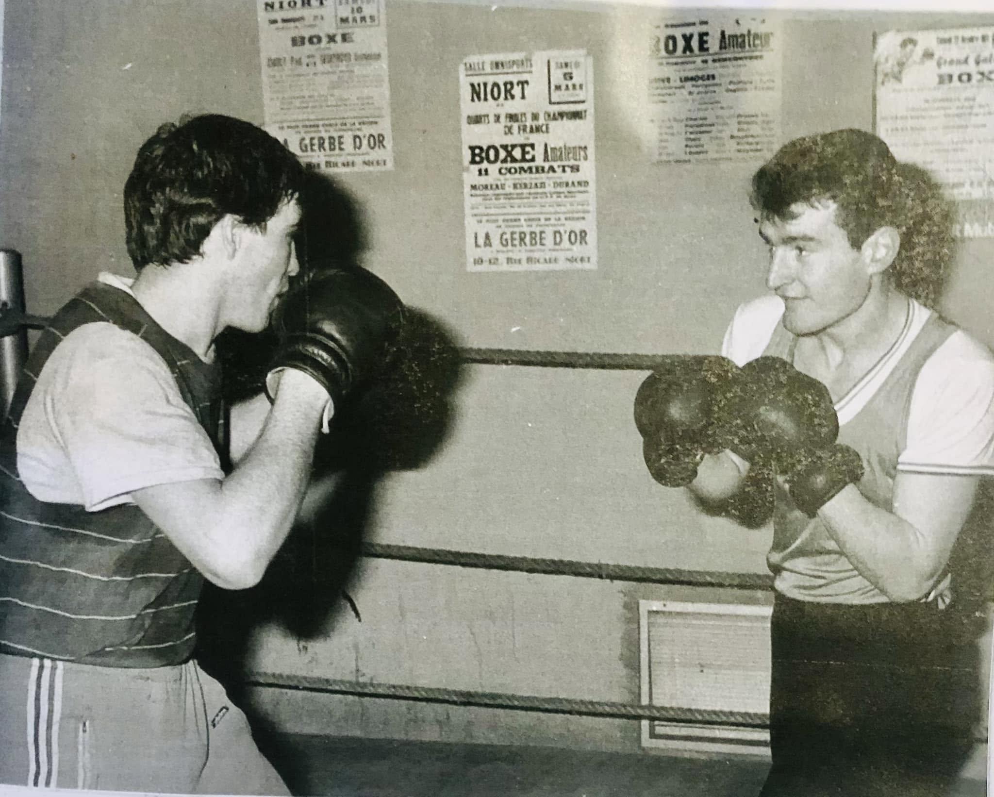Deux boxeurs en position dans un ring, affiches de combats amateurs en arrière-plan